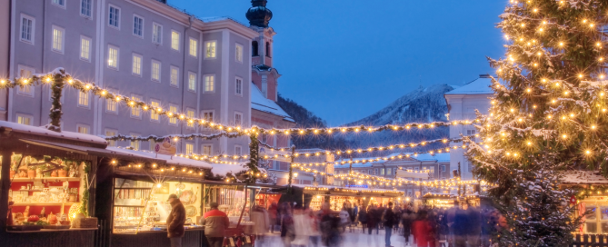 Twinkling Christmas market in a snowy European town at dusk, with warm lights, alpine scenery, and people exploring the festive stalls.